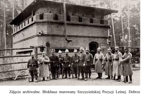 Żołnierze pozują przed murowanym blokhauzem, jednym z bunkrów Hitlera, z widocznymi stanowiskami strzeleckimi.