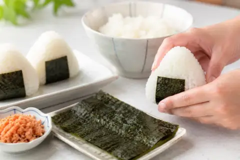 Dłonie formują pyszne onigiri. Obok miseczka z ryżem, nori i posypką.