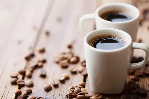 Dwa białe kubki z aromatycznym espresso na drewnianym blacie, otoczone ziarnami kawy. Zastanawiasz się, ile mg kofeiny ma espresso?