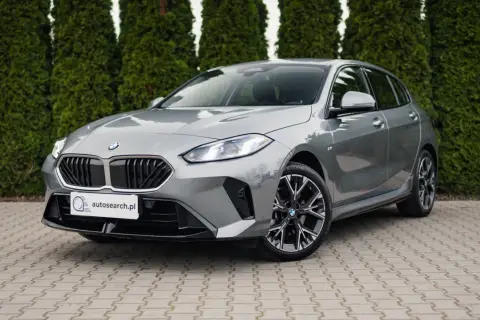 Szare BMW 118d z czarnym grillem i felgami stoi przed żywopłotem.