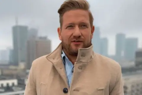 Bartek Jędrzejak w płaszczu na tle miasta. Marzy o powrocie do żony i dzieci po pracy.