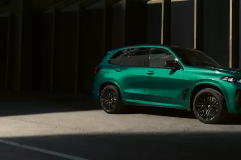 Zielony BMW X5 M z czarnymi felgami stoi na tle ciemnej architektury, oświetlony pasem światła.