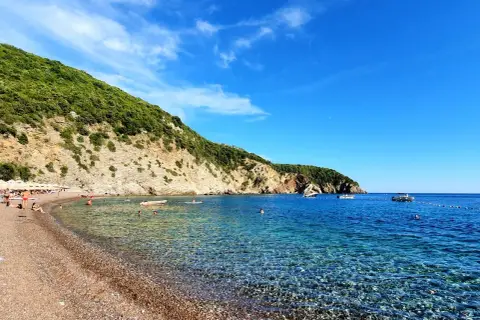 Kamienista plaża w Czarnogórze z turkusową wodą i zielonym wzgórzem w tle.
