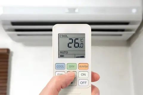 Dłoń naciska przycisk na pilocie klimatyzacji, wybierając funkcje chłodzenia. Na ekranie widoczna temperatura 26°C.