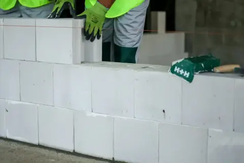 Budowa ściany z białych pustaków silikatowych. Ręce w zielonych rękawicach układają kolejny blok.