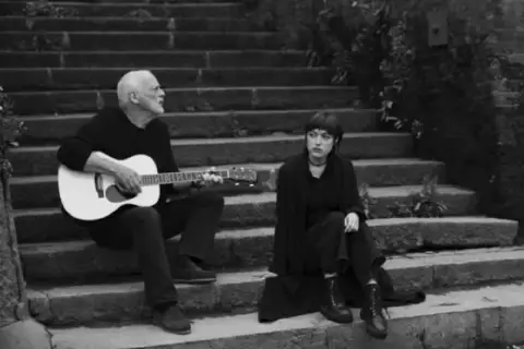 David Gilmour gra na gitarze, a obok niego siedzi jego córka.