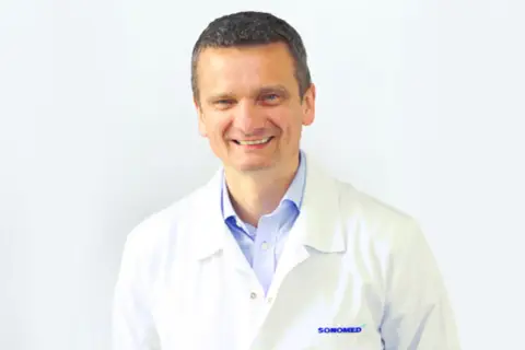 Uśmiechnięty mężczyzna w białym fartuchu lekarskim na jasnym tle.