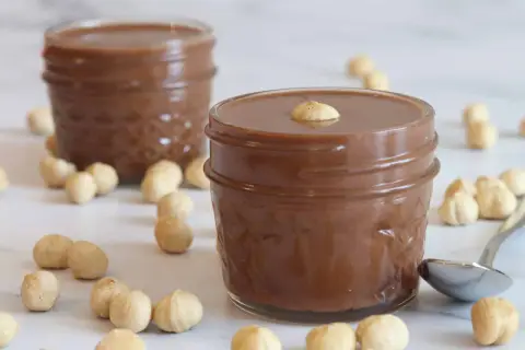 Dos tarros de deliciosa nutella vegana rodeados de avellanas. Una cucharilla plateada descansa cerca.