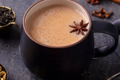 Aromaticzny napój, idealny na chłodne dni. Gorący **masala chai** w czarnej filiżance, ozdobiony anyżem. Obok liście herbaty, kardamon i goździki.