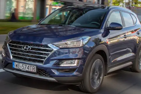 Nowy Hyundai Tucson w ciemnoniebieskim kolorze jedzie ulicą. Elegancka krata wlotu powietrza i nowoczesne reflektory podkreślają jego dynamiczny wygląd.