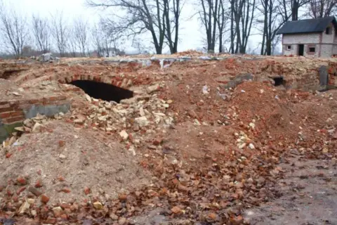 Ruiny ceglane z łukiem i mały domek w tle, otoczone drzewami.