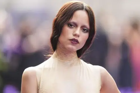 Jenna Ortega, czy ma polskie korzenie? Aktorka w kremowej sukni z siateczkowym kołnierzem, z burzą brązowych włosów i wyrazistym makijażem.