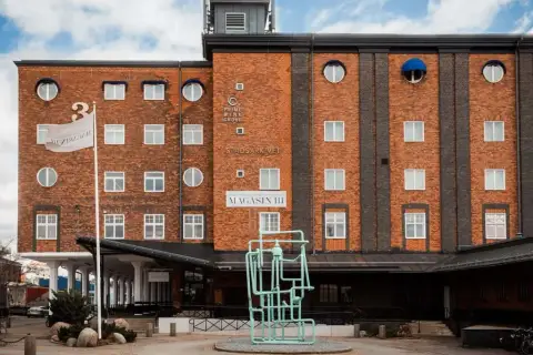 Magasin III, en byggnad i tegel med många fönster, har en flagga med texten "Magasin III". Framför finns en grön skulptur.