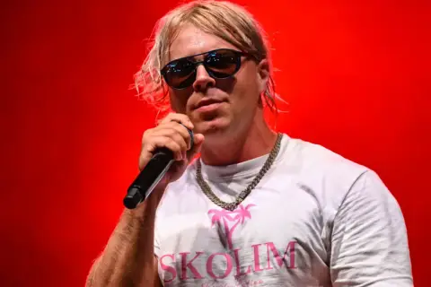 Skolim w okularach i białej koszulce z napisem "SKOLIM" śpiewa na czerwonym tle. Zastanawiasz się, ile Skolim bierze za koncert?