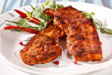 Dwa soczyste kawałki boczku z grilla, przyprawione i udekorowane papryczkami chili i zieleniną.