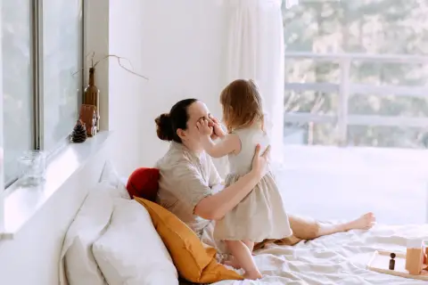 Dziecko dotyka nosa mamy, uosabiając wychowanie bliskościowe. Czuły moment na łóżku, otoczonym jasnymi poduszkami i światłem.