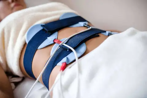 Osoba leży pod kocem z pasami i elektrodami na brzuchu, przygotowana do zabiegu ems trening.
