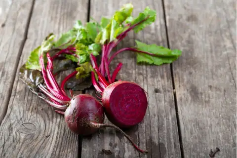 Zwei frische rote Bete, eine davon angeschnitten, liegen mit grünen Blättern auf Holz. Perfekt, um rote Beete roh zu essen.