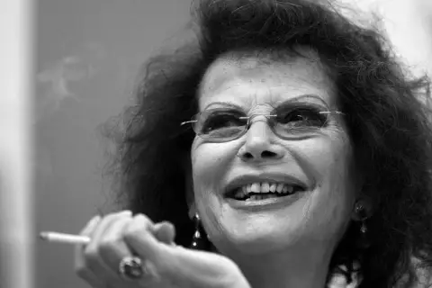 Uśmiechnięta Claudia Cardinale, ikona kina, z papierosem w dłoni. Jej spojrzenie pełne jest życia, jak w jej najlepszych filmach.