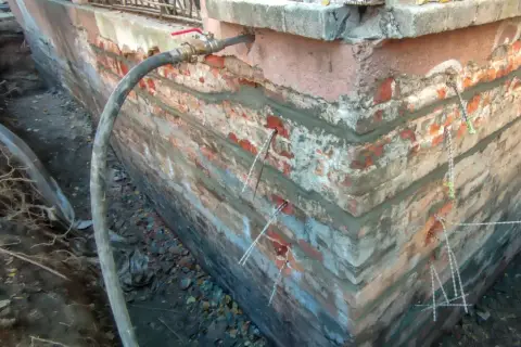 Naprawa pęknięć i osiadania budynku. W ścianie fundamentowej widoczne są kotwy wzmacniające i wąż doprowadzający zaprawę.