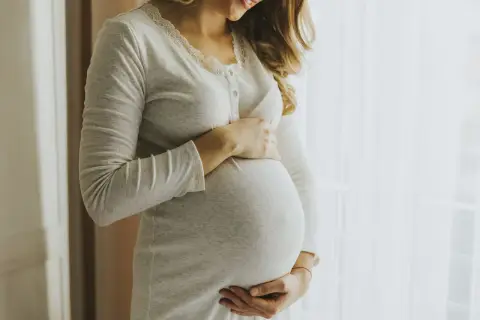 Kobieta w ciąży, z dłońmi na brzuchu, wygląda na szczęśliwą. Jej uśmiech sugeruje, że jest pełna życia, jak nimfomanka.