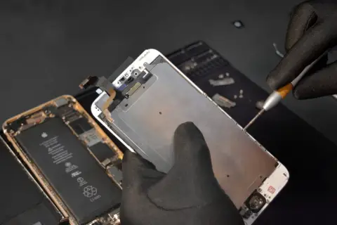 Ręka w czarnej rękawiczce przykręca śrubkę w rozebranym iPhonie.
