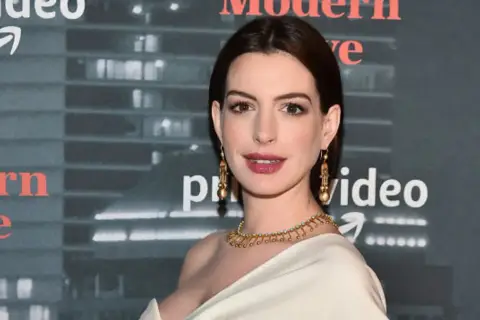 Anne Hathaway, której wiek jest często przedmiotem dyskusji, prezentuje się olśniewająco w złotej biżuterii na tle logo Amazon Prime Video.