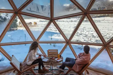 Pareja disfruta de un paisaje nevado en el Pirineo francés desde una cúpula geodésica. Vacas pastan en la distancia.