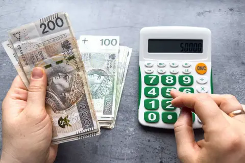 Ręka trzyma polskie banknoty 200 zł i 100 zł, obok kalkulator z wyświetloną kwotą 50000. Obliczenia związane z minimalną pensją.