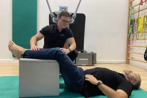 Fizjoterapeuta pomaga pacjentowi w ćwiczeniach na rwę kulszową. Pacjent leży na macie, z nogą opartą na podwyższeniu, a terapeuta wykonuje palpacje.