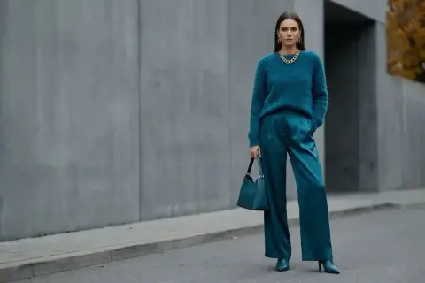 Elegancka kobieta w stylizacji w kolorze petrol, z błyszczącymi spodniami i miękkim swetrem, pozuje na tle betonowej ściany.