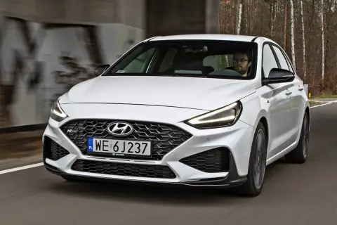 Biały Hyundai i30 N z dynamicznym grillem i agresywnym zderzakiem jedzie po drodze wśród drzew.