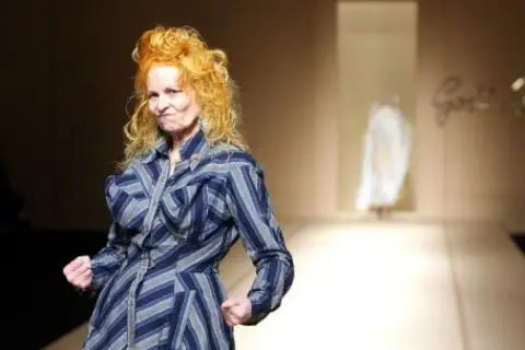 Vivienne Westwood, con su icónico cabello naranja, posa con energía en la pasarela de Westwood, luciendo un vestido a rayas azules y grises.