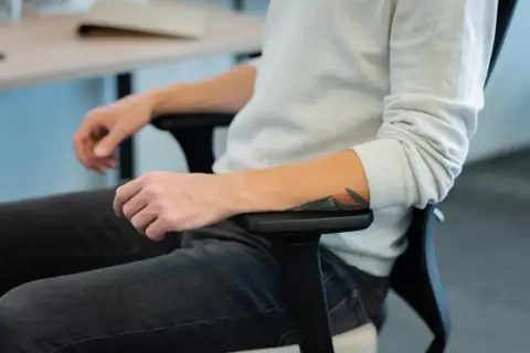 Osoba z tatuażem na ramieniu siedzi w biurowym fotelu, co może symbolizować siedzący tryb życia.