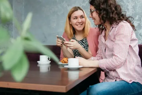 Dwie kobiety rozmawiają przy kawie, tworząc intymne tête-à-tête. Jedna pokazuje coś na telefonie, druga uśmiecha się.