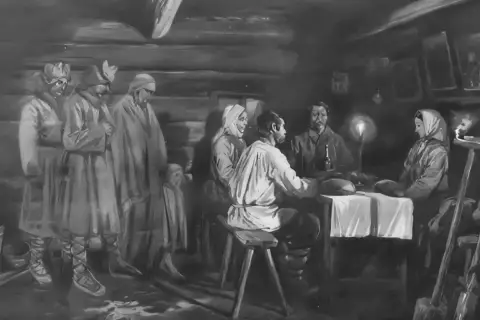 Ludzie w tradycyjnych strojach, w tym dzieci, zebrani w izbie. Na stole świeca, obok narzędzia. Atmosfera przypomina obrzędy dziadów.