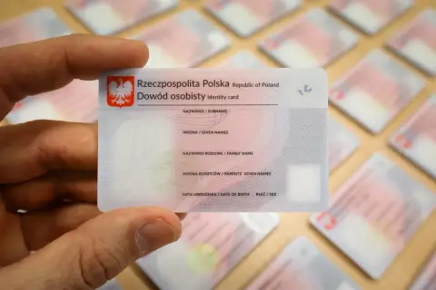 Dłoń trzyma polski dowód osobisty, jeden z wielu leżących na stole. To ważny dokument, zawierający Twój osobisty numer identyfikacyjny.