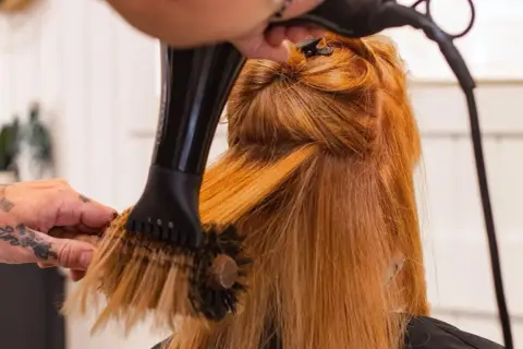 Manos con tatuajes secan y peinan cabello pelirrojo con secador y cepillo redondo. El brushing que es, es una técnica de estilismo.