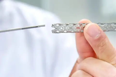 Dłoń trzyma metalowy stent o ażurowej strukturze, obok widoczny jest instrument medyczny.