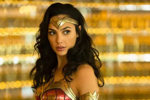 Gal Gadot jako Wonder Woman, część obsady filmu, w złotej zbroi i opasce na głowie, na tle rozmytych, złotych świateł.