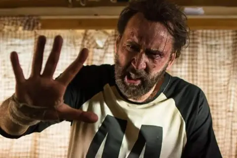 Nicolas Cage med blåmärken och svettig panna, höjer handen i en gest av stopp. En scen från en av hans filmer.