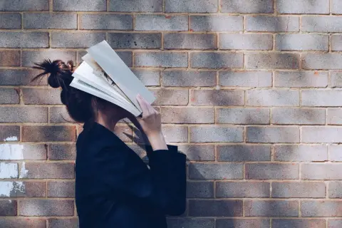 Persona con un libro abierto cubriendo su rostro, apoyada contra una pared de ladrillos. Parece estar inmersa en la lectura, quizás buscando un momento de tranquilidad.