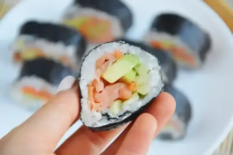 Ręka trzyma kawałek futomaka z łososiem i awokado. Na talerzu widać więcej sushi.
