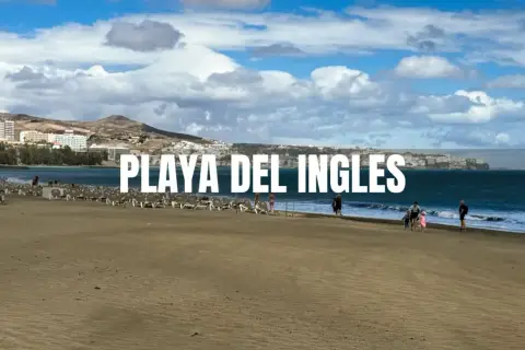 Piękna plaża Playa del Inglés z rzędami leżaków i kilkoma spacerującymi ludźmi. W tle widać hotele i góry.
