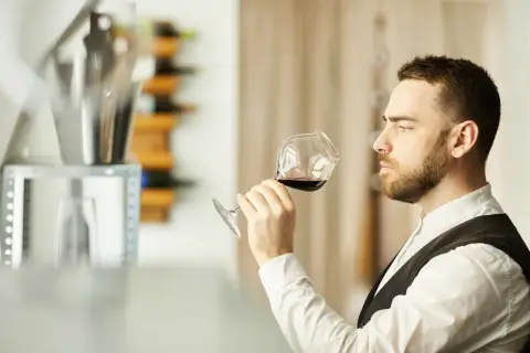 Mężczyzna w białej koszuli i kamizelce, sommelier, kto to? Wącha kieliszek czerwonego wina, oceniając jego aromat.