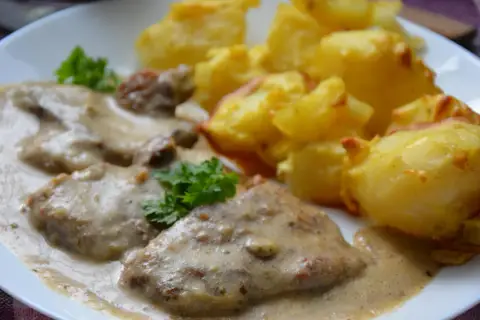 Pyszny karczek w sosie chrzanowym z pieczonymi ziemniakami. Danie ozdobione natką pietruszki.