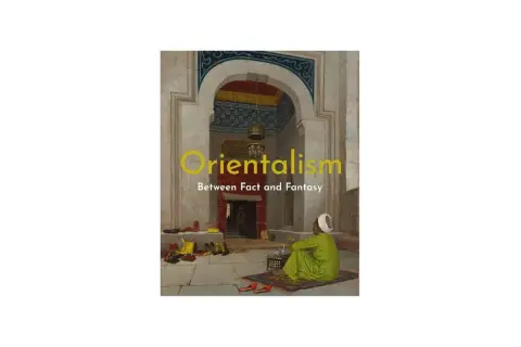 A man in a green robe sits on a rug in an ornate archway, a scene evoking orientalism art. Shoes are neatly arranged on the stone floor.