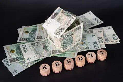Dom z banknotów 100 zł, symbolizujący kredyt hipoteczny. Słowo "KREDYT" ułożone z drewnianych pieczątek.