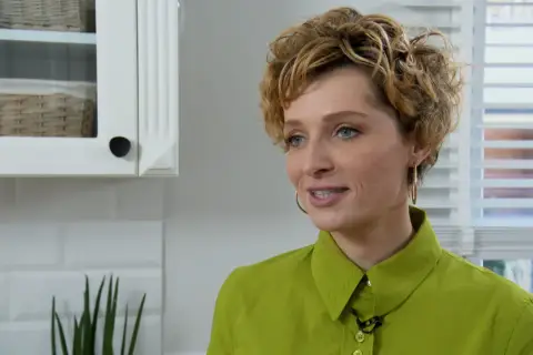 Katarzyna Jaroszyńska, wiek nieznany, w zielonej koszuli, z kręconymi blond włosami, uśmiecha się. W tle biała kuchnia.