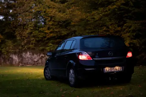 Czarny Opel Astra H 1.6 benzyna zaparkowany na trawie, w tle jesienne drzewa.